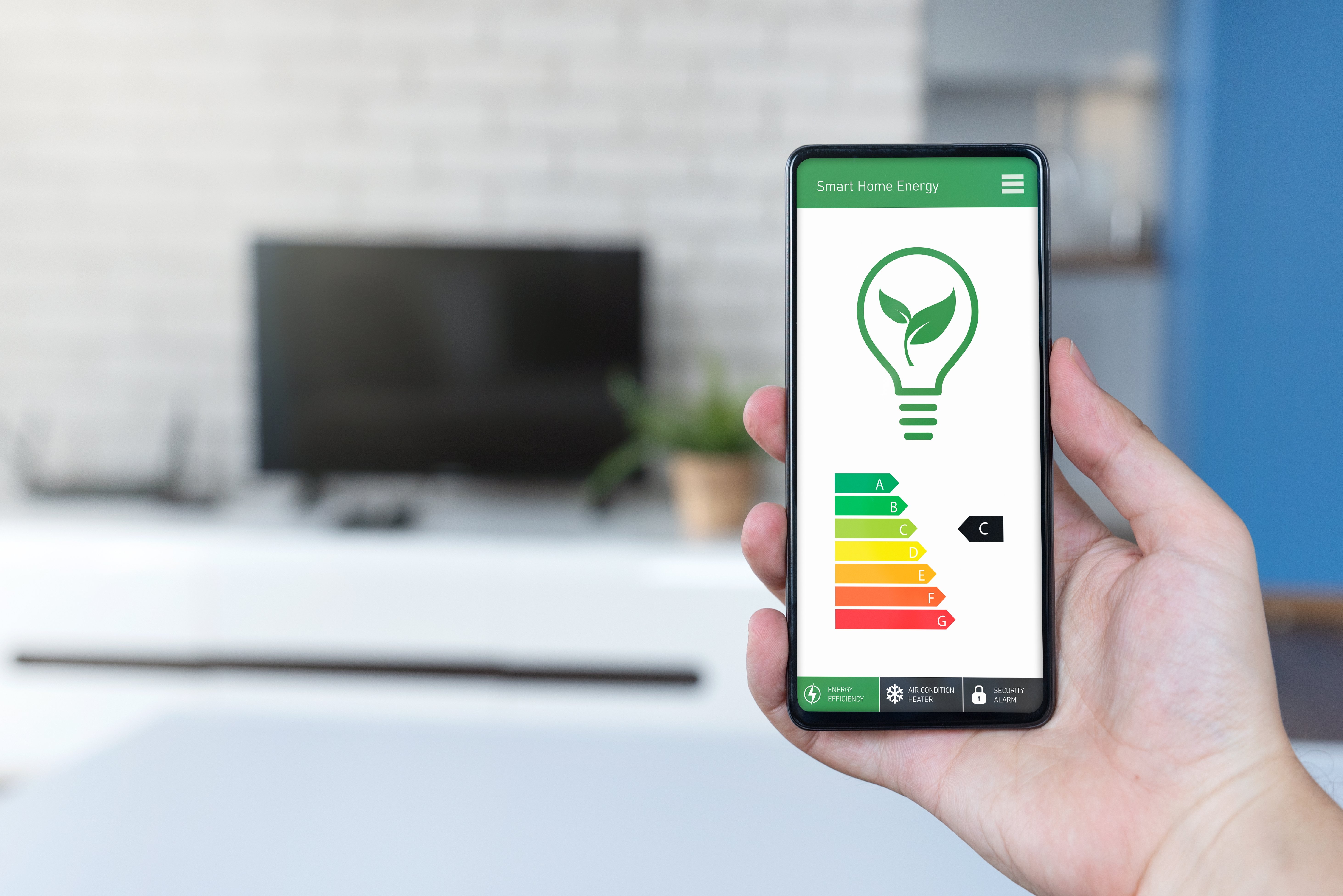 Eine Hand mit Handy auf dem die Energieklassen und eine Glühbirne dargestellt sind. Im Hintergrund sieht man verschwommen einen Fernseher.
