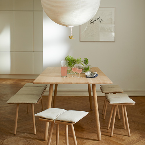 An einem Esstisch aus hellem Holz stehen drei Hocker und eine Bank im selben schlichten Stil mit beigen Sitzkissen. Über dem Tisch hängt eine große, weiße, kugelförmige Papierleuchte.