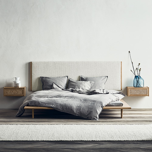 Ein niedriges Futonbett mit Bettwäsche aus grauem Leinen. Das Kopfteil ist ebenfalls mit hellgrauem Stoff bezogen. Links und rechts neben dem Bett befinden sich kleine Hängeschränke mit Fronten aus Wiener Geflecht als Nachttische.
