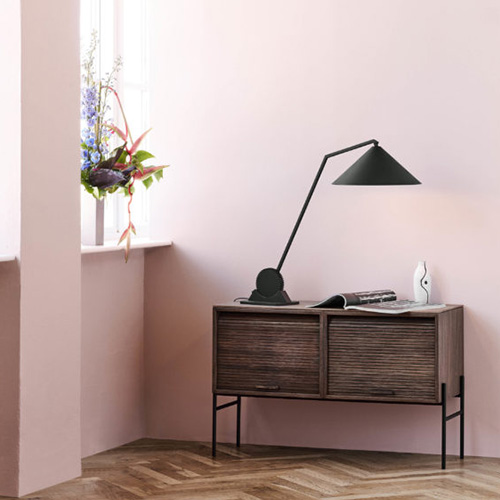 Ein Sideboard aus gebeiztem Holz steht auf filigranen schwarzen Metallfüßen. Die Fronten der Schiebetüren sind aus Holzlamellen. Auf dem Sideboard steht eine grafisch gestaltete Tischleuchte aus schwarzem Metall.