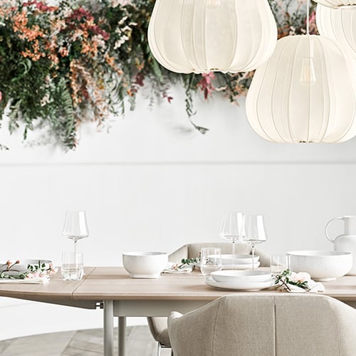 Über einem hellen Holztisch gedeckt mit weißem Geschirr, hängen mehrere mit weißen transluzentem Stoff bezogene Hängeleuchten. Die weiße Wand ist mit bunten Blumen und grünem Farn geschmückt. Am Tisch stehen schlichte Polsterstühle mit cremefarbenem Bezug.