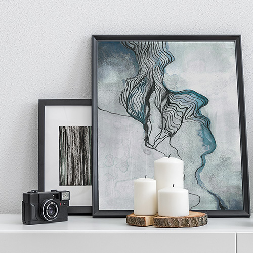 Auf einem weißen Sideboard stehen zwei an die Wand gelehnte abstrakte Bilder mit schwarzem Rahmen. Davor sind drei weiße Kerzen auf einer naturbelassenen Holzscheibe dekoriert.